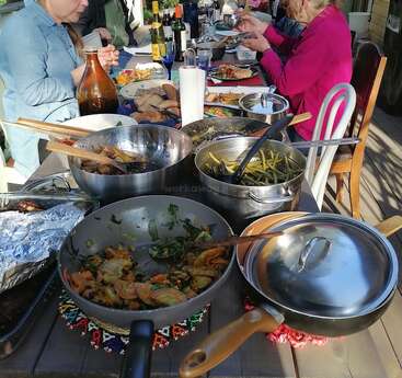 The image depicts a table laden with an assortment of food and cooking vessels, surrounded by individuals seated at the table, enjoying their meal.