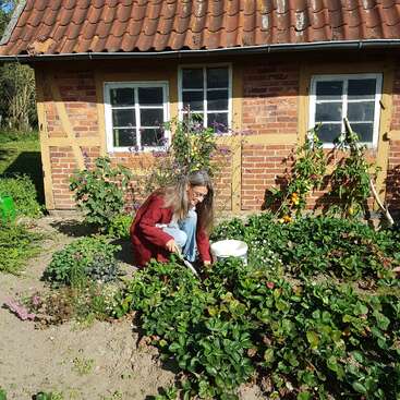 Une femme aux cheveux longs s'occupe d'un jardin devant une petite maison en briques au toit de tuiles, par une journée ensoleillée.