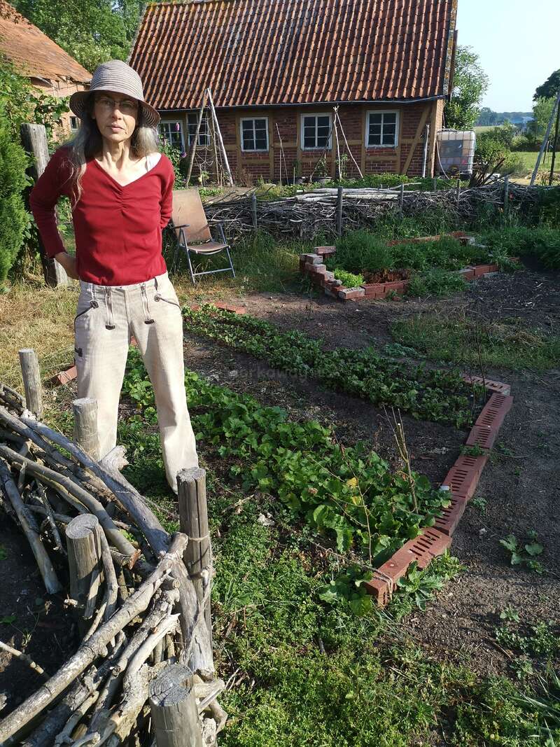 A imagem mostra uma mulher em um jardim, vestindo camisa vermelha e calça bege, com uma casa rústica e vegetação exuberante ao fundo em um dia ensolarado.
