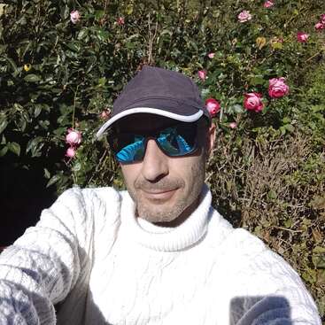 Un homme vêtu d'un pull blanc et de lunettes de soleil bleues réfléchissantes prend un selfie en plein air. Il porte une casquette, entouré de buissons verts et de roses roses en fleur.