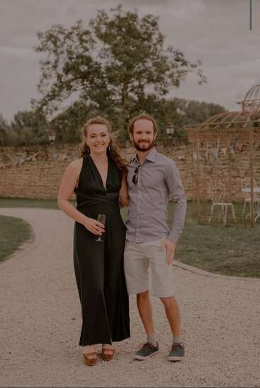 A smiling couple poses outdoors on a gravel path. The woman in a black dress holds a drink, while the man wears a shirt and shorts. Trees background.