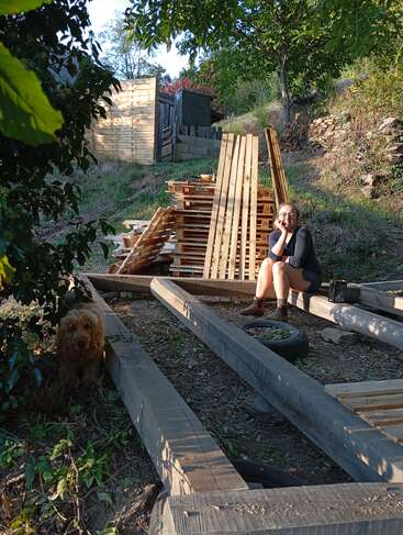 A woman sits smiling on wooden beams near stacked planks outdoors, surrounded by greenery. A brown dog is partially hidden in the shade, watching quietly.