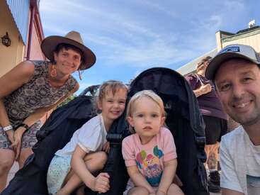 Eine lächelnde vierköpfige Familie posiert an einem sonnigen Tag für ein Foto im Freien. Zwei kleine Kinder sitzen in einem Kinderwagen, flankiert von ihren Eltern.