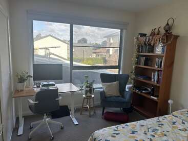 A cozy bedroom workspace with a wooden desk, office chair, computer, armchair, bookshelf, indoor plants, large window, and bed with a floral-patterned duvet.