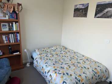 A cozy bedroom features a bed with floral bedding, a wooden bookshelf, a small lamp, two nature photos on the wall, and minimalistic, tidy decor.