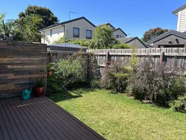 A sunny backyard with a wooden deck, green lawn, lush plants along the fence, potted plants, and a green watering can. Suburban houses in background.
