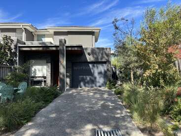 A modern two-story house with a black brick facade, single garage, wide driveway, lush green garden, and outdoor seating area under a clear blue sky.