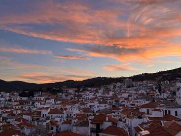 White-washed houses with red-tiled roofs cluster on a hillside beneath a dramatic sunset sky, warm orange and pink clouds stretching above a tranquil Mediterranean village scene.