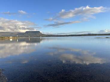Un lac paisible reflète un ciel bleu et des nuages épars, avec une montagne lointaine au sommet plat et des collines vertes sous une lumière du jour éclatante, créant un paysage naturel et serein.