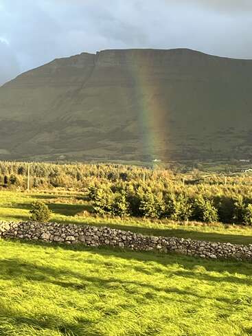 Ein leuchtender Regenbogen fällt auf ein üppiges grünes Feld, das von einer Steinmauer begrenzt wird, mit einem Berg und einem dramatischen Himmel im ruhigen Hintergrund. Friedliche Landschaft.
