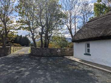 Une petite maison pittoresque au toit de chaume se trouve au bord d'une allée de gravier. Des murs de pierre bordent l'entrée, et de grands arbres verts projettent des ombres pommelées dans la lumière du jour.