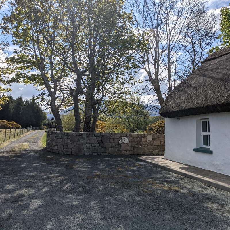 Une petite maison pittoresque au toit de chaume se trouve au bord d'une allée de gravier. Des murs de pierre bordent l'entrée, et de grands arbres verts projettent des ombres pommelées dans la lumière du jour.