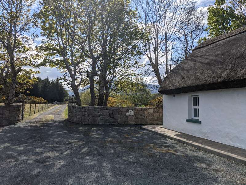 Ein malerisches, strohgedecktes Häuschen steht neben einer Schotterauffahrt. Steinmauern säumen die Einfahrt und hohe grüne Bäume werfen im hellen Tageslicht schummrige Schatten.