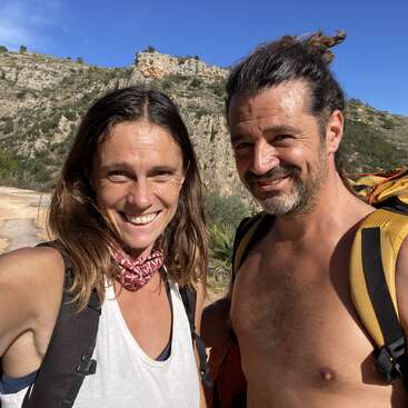 Une femme et un homme souriants posent à l'extérieur par temps ensoleillé, portant des sacs à dos. Des collines rocheuses et un ciel bleu clair forment la toile de fond, suggérant une randonnée ou un voyage d'aventure.
