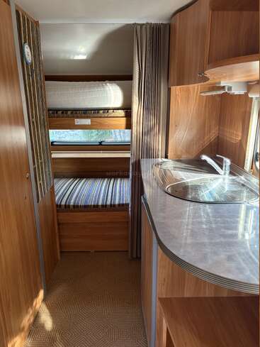 Cette image montre l'intérieur d'un camping-car compact avec un décor en bois, des lits superposés, une petite fenêtre, une kitchenette avec un évier et une moquette à motifs.