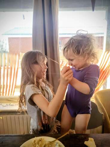 Dos niños pequeños juegan alegremente juntos en casa, junto a una ventana. Uno está subido a una silla; ambos se ríen y se dan la mano, y en la mesa se ve comida y tarta.