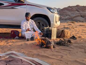 Ein Mann in traditioneller Kleidung sitzt an einem Lagerfeuer in der Wüste und bereitet Tee zu. In der Nähe ist ein weißes Fahrzeug geparkt und im Hintergrund sind die Berge zu sehen.
