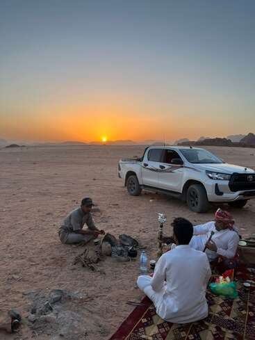 Drei Männer sitzen bei Sonnenuntergang auf einem Teppich in der Wüste, entspannen sich neben einem weißen Lastwagen, trinken Tee und rauchen Shisha und genießen einen friedlichen Abend.