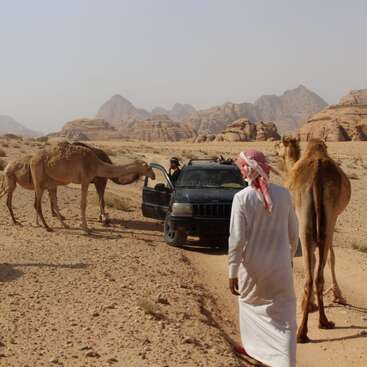 A man stands in front of a large body of water, wearing a collared shirt and pants, with a serious expression on his face. The background is a body of water.