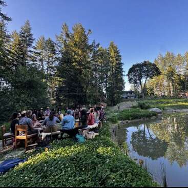 A group of people gather outdoors in a lush, green area beside a pond, surrounded by tall trees under a clear blue sky, enjoying nature together.