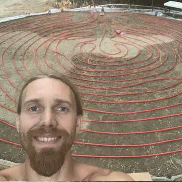 Um homem sorridente tira uma selfie em frente a um grande e complexo padrão de labirinto feito de tubos vermelhos dispostos em um terreno seco e marrom ao ar livre.