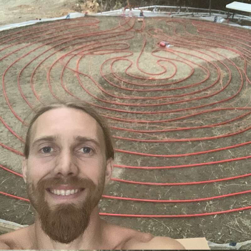 A smiling man takes a selfie in front of a large, complex labyrinth pattern made from red tubing or pipe arranged on dry, brown ground outdoors.