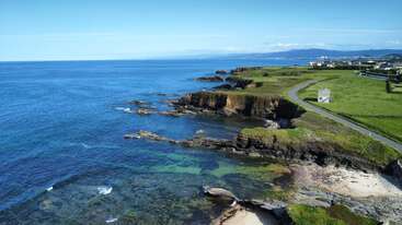 A beautiful coastline with clear blue water, rugged cliffs, and green fields. A winding road follows the shore, leading past a small building under a vast sky.