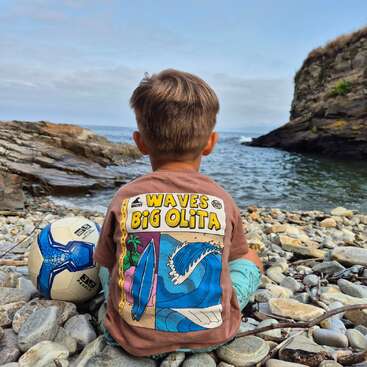 A young boy sits on rocky shore, facing the sea. Wearing a colorful "Waves Big Olita" shirt, a soccer ball beside him, he enjoys the peaceful coastline.