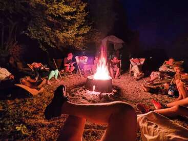 Das Bild zeigt eine Gruppe von Menschen, die nachts um ein Lagerfeuer versammelt sind. Einige sitzen auf Stühlen, andere liegen auf dem Boden und genießen die warme Glut.