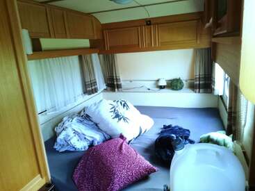 Cette image montre une chambre de camping-car confortable avec des armoires en bois, un grand lit avec des oreillers et de la literie, des petits rideaux aux fenêtres et une cruche d'eau à proximité.