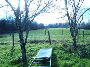 Une chaise longue est posée sur l'herbe verte entre deux arbres sans feuilles. Une clôture en bois entoure le champ ouvert, avec une zone forestière à l'arrière-plan. Le soleil brille.