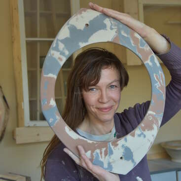 The image depicts a woman holding a large, circular ceramic piece with a hole in the center, showcasing her artistic creation in a workshop setting.