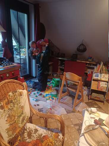 Un momento alegre en casa: un adulto levanta a un niño en un gesto juguetón por encima de unos juguetes de colores. Una habitación acogedora y llena de cosas, con libros, sillas y un ambiente alegre.