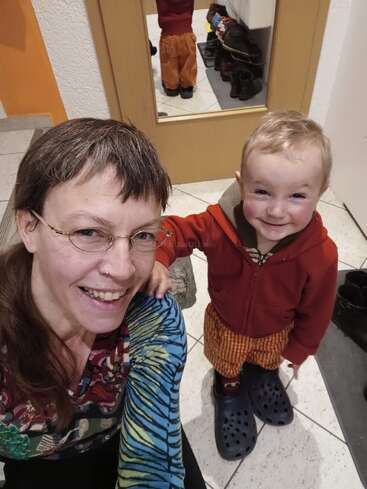 Una mujer sonriente se hace un selfi con un niño feliz que lleva unos zapatos demasiado grandes. Ambos están en el interior, con un espejo detrás de ellos en el que se refleja la espalda del niño. Un momento acogedor y alegre.