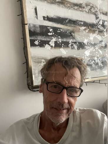 A man with glasses and tousled hair is sitting indoors, smiling gently. Behind him is a textured, abstract black-and-white painting framed on the wall.