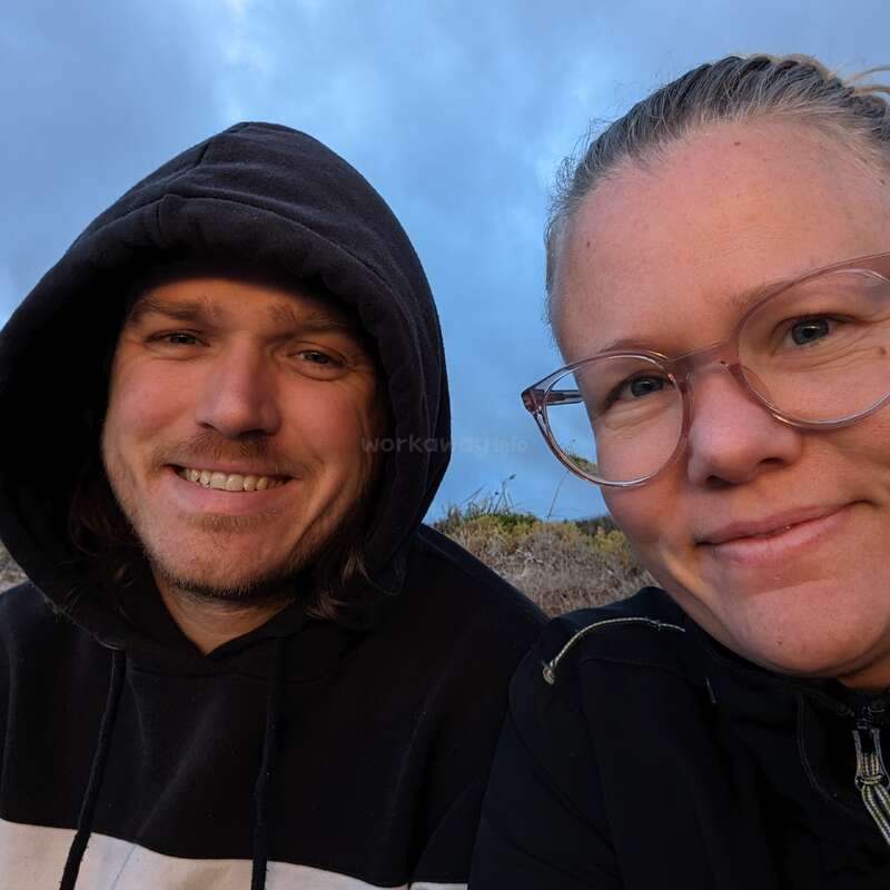 A smiling man in a black hoodie and a woman wearing glasses and a black jacket, outdoors under a cloudy sky, enjoy a relaxed moment together.