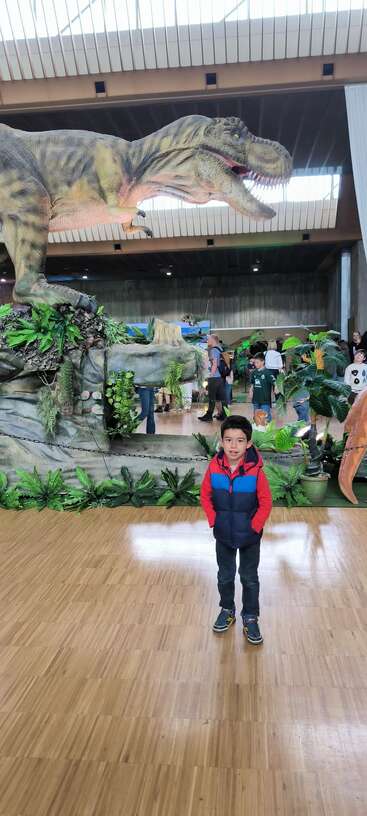 Un jeune garçon se tient à l'intérieur et sourit à l'appareil photo. Derrière lui se trouve une grande maquette de dinosaure très réaliste, entourée de plantes artificielles et d'autres visiteurs du musée.