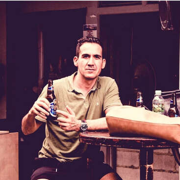 Un homme est assis à une table ronde en plein air, une bière à la main. Il porte une chemise verte, fait un geste de la main et regarde directement la caméra.