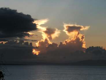Das Bild zeigt einen heiteren Sonnenuntergang über einem Gewässer, mit großen Wolken, die von den Sonnenstrahlen beleuchtet werden, und einer dunklen Wolke auf der linken Seite.