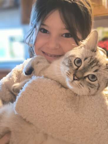 Un niño sonriente abraza con cariño a un gato mullido y de pelaje claro. El gato parece tranquilo, pero curioso, y mira fijamente a la cámara. Una luz cálida y suave inunda esta acogedora escena.