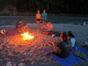 Un grupo de amigos se sienta alrededor de una hoguera en la orilla arenosa de un río al atardecer, charlando, relajándose y disfrutando juntos del calor y del ambiente tranquilo al aire libre.