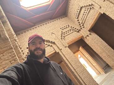 Um homem barbudo, usando um boné marrom e um moletom preto com capuz, está dentro de um prédio rústico com alvenaria decorativa e luz natural iluminando o espaço.