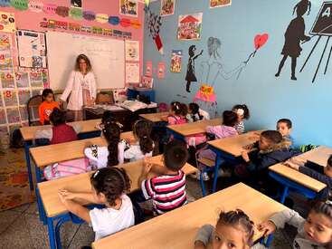 Uma sala de aula decorada com cores vivas, com crianças pequenas sentadas nas carteiras, olhando atentamente para uma professora sorridente perto do quadro branco, rodeadas por pôsteres coloridos e decorações nas paredes.