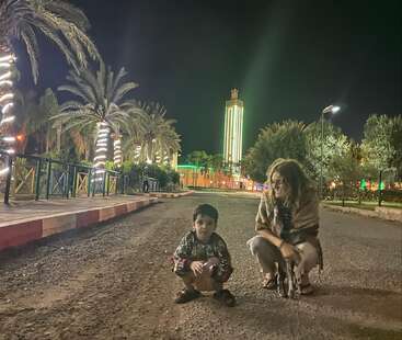 Uma mulher e uma criança estão agachadas numa rua da cidade à noite, rodeadas por palmeiras enfeitadas com luzes. Edifícios iluminados por néon brilham ao fundo.