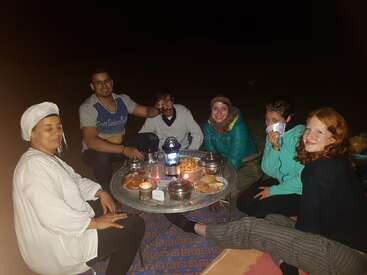 Cinco pessoas estão sentadas ao redor de uma mesa baixa ao ar livre, à noite, compartilhando comida e bebida. Elas parecem felizes, relaxadas e estão curtindo a companhia umas das outras, jogando cartas.