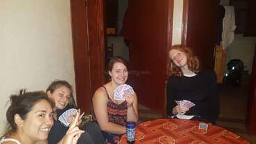 Quatro jovens estão sentadas ao redor de uma mesinha jogando cartas, sorridentes e descontraídas. O cenário parece ser um ambiente interno, com iluminação aconchegante e um clima descontraído.