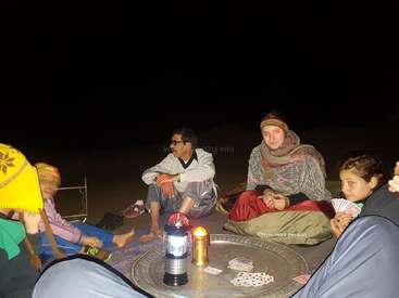 Um grupo de pessoas está sentado no chão ao redor de uma mesa ao ar livre à noite, jogando cartas à luz de uma lanterna, vestidas com roupas quentes e parecendo relaxadas e alegres.
