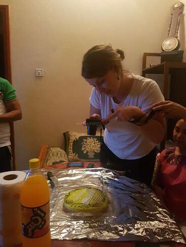 Uma mulher está tirando uma foto de um bolo decorado em cima de uma mesa coberta com papel-alumínio. Perto dali estão uma garrafa de refrigerante de laranja, toalhas de papel e outras pessoas.