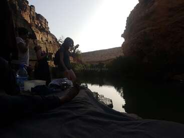 Um grupo de pessoas relaxa à beira de um rio calmo, cercado por penhascos rochosos, ao pôr do sol; algumas se preparam para dar um mergulho, com suas silhuetas recortadas contra o céu incandescente.