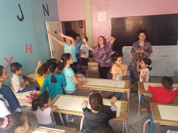 A imagem mostra uma sala de aula animada com crianças e professores envolvidos em uma atividade, cercados por paredes e carteiras coloridas, transmitindo uma sensação de alegria e aprendizado.
